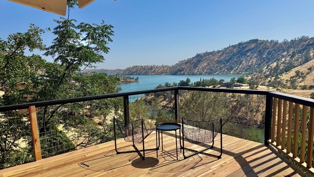 A wooden deck with a metal railing, two chairs, and a view of a lake and hills. - Lake Haus 59 : Family-friendly 2bed/2ba, Full Kitchen + Lake View Deck (Friant)