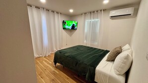 Ein Schlafzimmer mit Bett, Vorhängen, einem an der Wand montierten Fernseher und einer Klimaanlage.