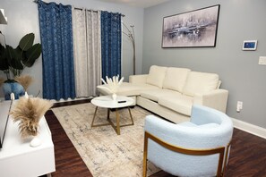 A living room with a white sofa, a round coffee table, a blue armchair, a large painting, and a plant. - 2 bedroom apartment in a quiet area of Newark, near the airport and stores. (Newark)