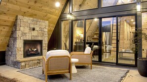 Un intérieur chaleureux avec une cheminée en pierre, des meubles en osier et une grande porte vitrée donnant sur un espace cuisine.