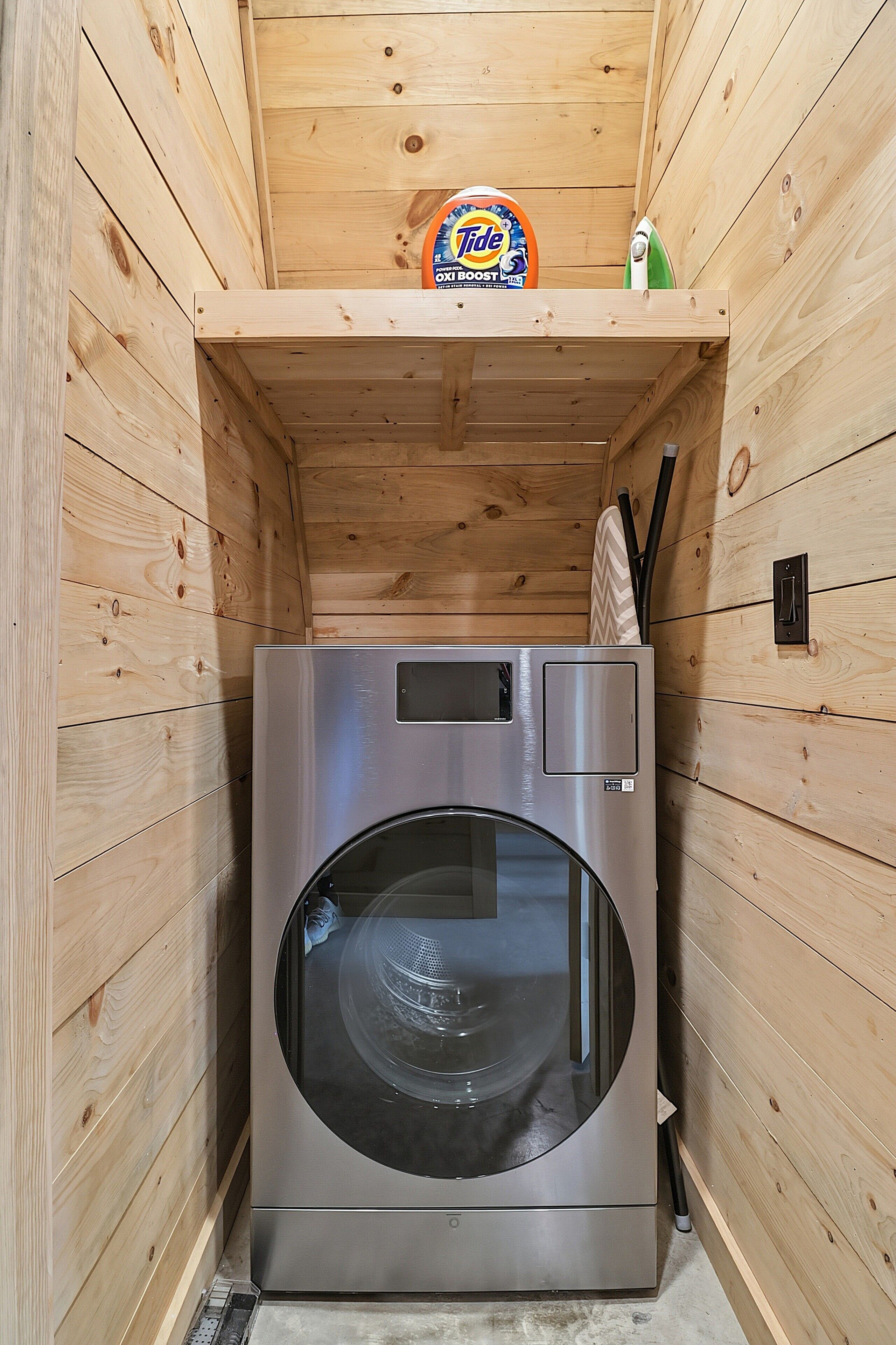 A compact, wooden-walled laundry room with a stainless steel washer and dryer, a Tide detergent box, and a green cleaning product on the shelf.