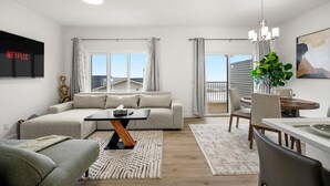 A modern living room with a sectional sofa, a dining table with chairs, a TV mounted on the wall, and a large window with curtains. - Mountain View Retreat with Two King Beds and Full House (Cochrane)