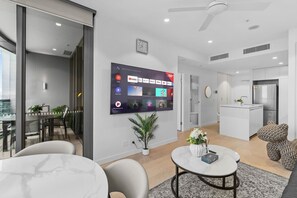 En moderne stue med et fladskærms-tv, et spiseområde med et glasbord og et køkken med indbygget køleskab.
