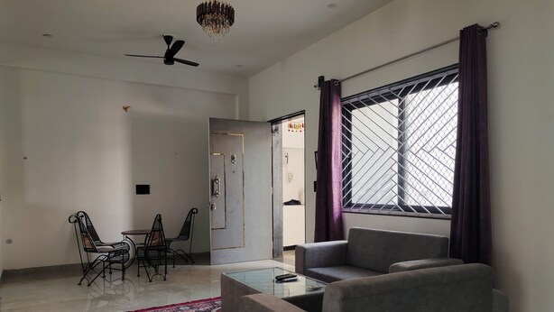 Ein Wohnzimmer mit grauem Sofa, einem Couchtisch, einem gemusterten Teppich, einem Essbereich mit Stühlen, einem Deckenventilator und einem Fenster mit Vorhängen.