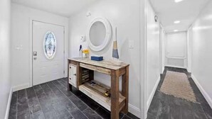 Een gang met een witte deur, een houten bijzettafeltje, een spiegel en een vloerkleed.