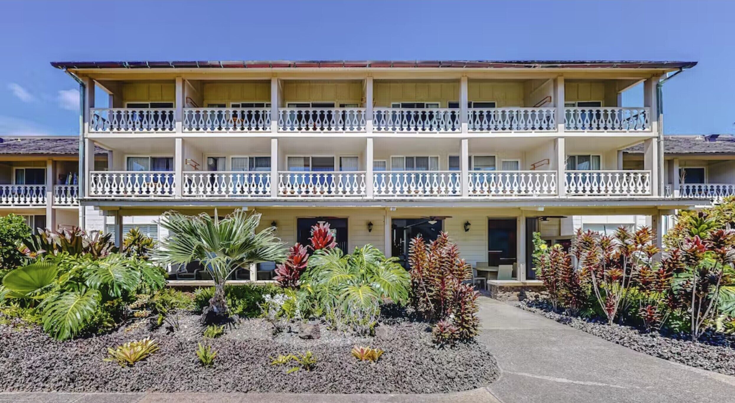 A two-story hotel with a balcony on each floor, a covered entrance, and tropical landscaping.