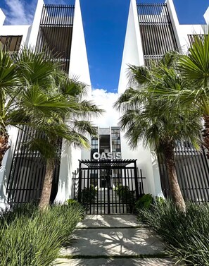 A modern building with a gated entrance, palm trees, and a clear blue sky. - Tropical Oasis in Bayahibe with Pool and Private Terrace with Jacuzzi and BBQ! (Bayahíbe)
