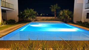 Una piscina rectangular con superficie de agua azul cristalina, rodeada de césped cuidado y palmeras, con edificios modernos al fondo.