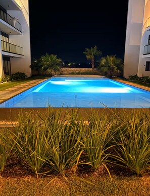 A rectangular swimming pool with a clear blue water surface, surrounded by well-maintained grass and palm trees, with modern buildings in the background. - Tropical Oasis in Bayahibe with Pool and Private Terrace with Jacuzzi and BBQ! (Bayahíbe)