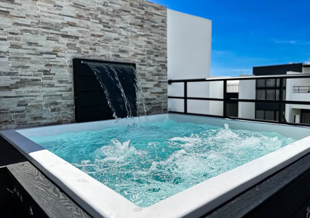 Un moderno jacuzzi al aire libre con caída de agua, frente a una pared de piedra y un cielo azul despejado.