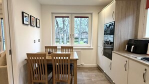 A modern kitchen with a dining area, featuring a wooden table and chairs, a built-in oven, and a window with a view of trees. - Ness Gardens, Inverness (Inverness)