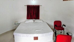 Uma cama de solteiro com colchão branco e cortinas vermelhas, uma cadeira vermelha e uma mesa de vidro com um objeto preto em cima.