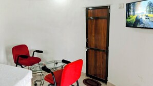 Um quarto com porta de madeira, mesa de vidro e cadeiras, e uma televisão na parede.