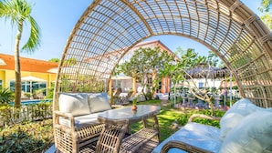 A covered outdoor seating area with wicker furniture and a glass table, surrounded by greenery and a pool. - Tongtong Garden Holiday Hotel (Rawai)