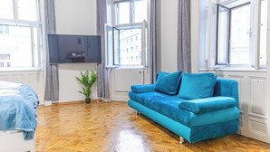 A modern living room with a blue sofa, a flat-screen TV, and large windows offering city views. - Comfy 2BR Apt For 6 Wiener Stadthalle (Vienna)