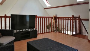 Amplia sala de estar con televisor de pantalla plana, sofá, mesita de centro y ventilador de techo. Se observan barandales de madera y una escalera que conduce a un nivel superior.