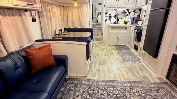 A compact RV interior with a blue sofa, a patterned rug, a kitchenette, and a ceiling fan.