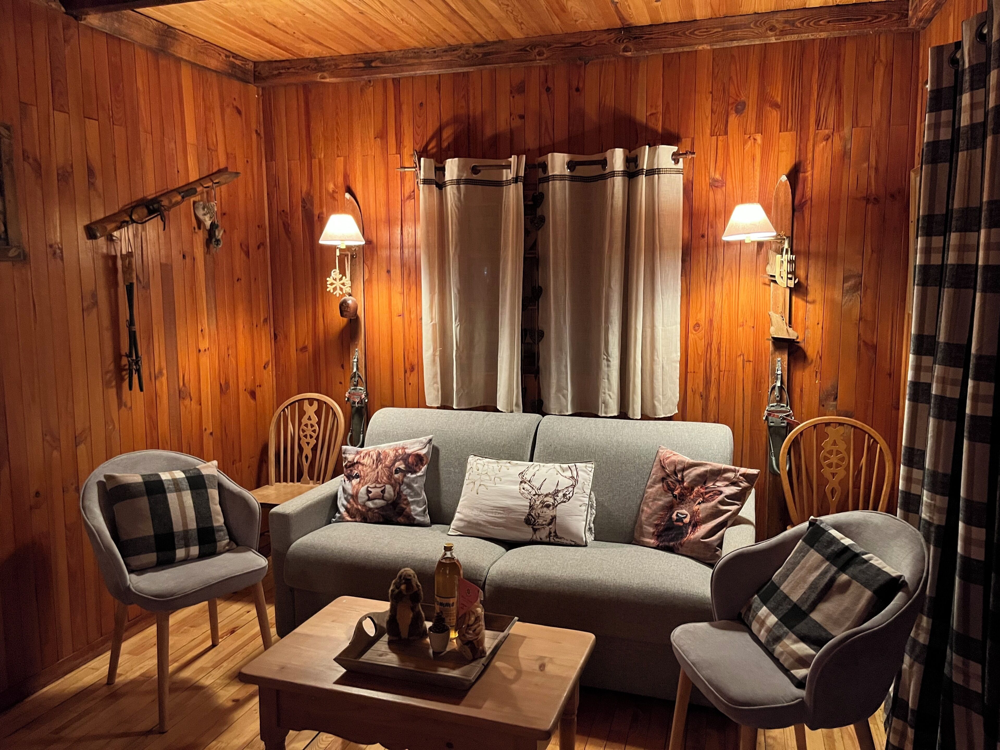 A cozy room with wooden walls, a grey sofa, two armchairs, a wooden coffee table, and a window with curtains.