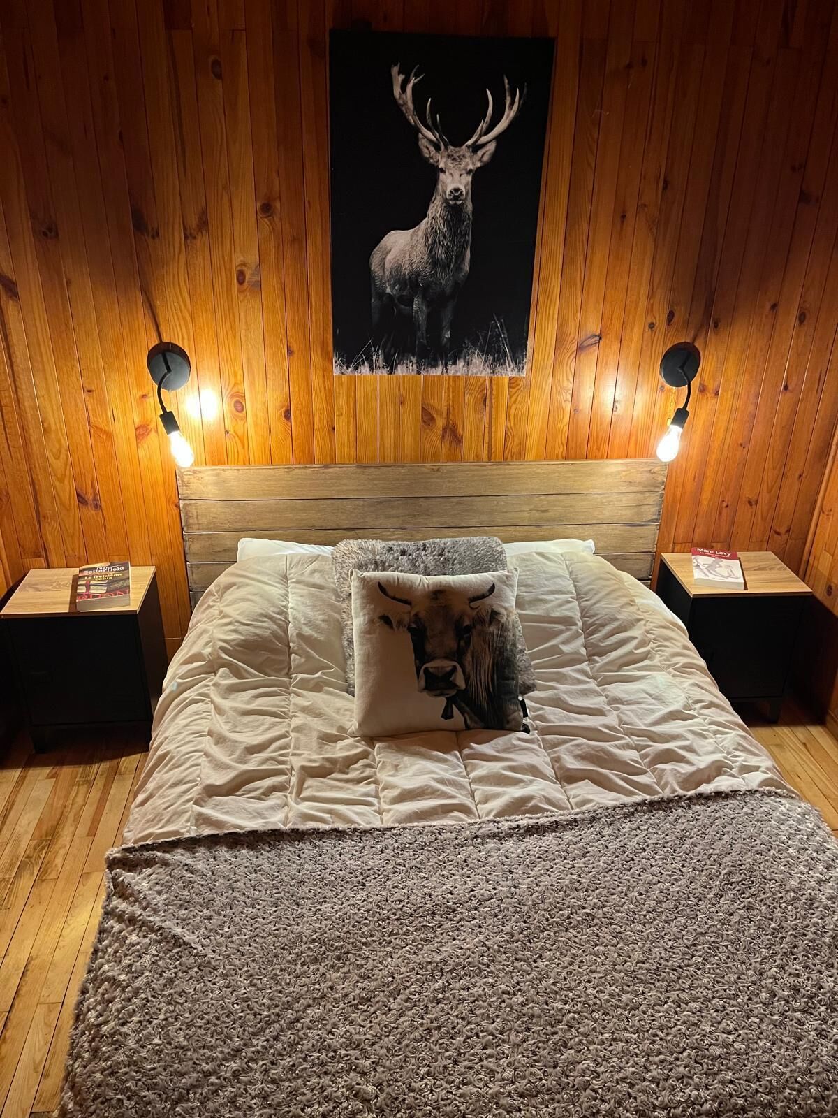 Une chambre à coucher avec une tête de lit en bois, un lit recouvert d’une couette blanche, d’une couverture grise et d’un oreiller orné d’un motif de vache. On y trouve deux tables de chevet avec des lampes et une affiche représentant un cerf accrochée au mur.