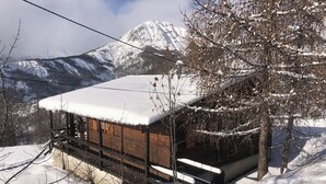 A wooden cabin with a snow-covered roof, surrounded by snow-covered trees and mountains in the background.