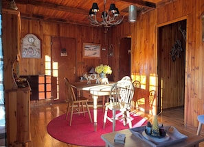 A wooden interior with a dining table, chairs, a chandelier, and a clock.