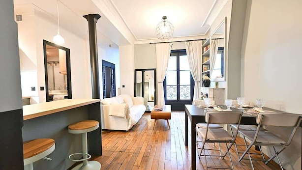 A modern kitchen with a wooden floor, white bar stools, a white sofa, and a dining area with metal chairs. - Élégant 2 chambres au cœur du Marais (Paris)