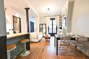 A modern kitchen with a wooden floor, white bar stools, a white sofa, and a dining area with metal chairs. - Élégant 2 chambres au cœur du Marais (Paris)