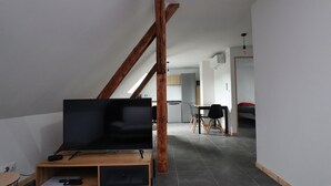Ein modernes Wohnzimmer mit Fernseher auf einem Holzständer, einem Essbereich mit Tisch und Stühlen sowie einer Küche im Hintergrund.