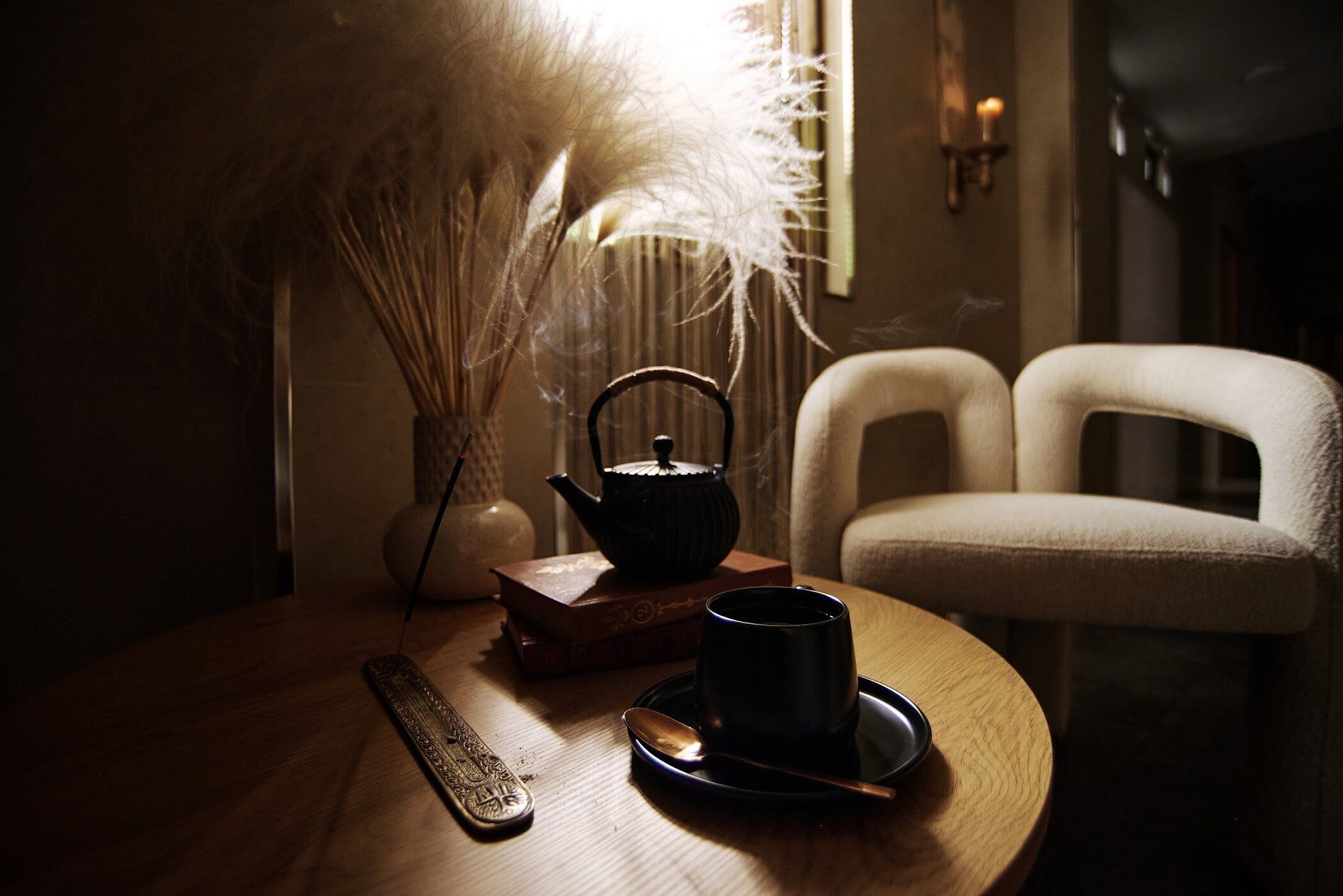 A round wooden table with a black teapot, a cup, and a spoon on a saucer. There is a book and a decorative vase with dried plants on the table. A chair is partially visible to the right.