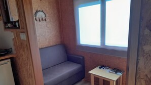 A small room with a wooden wall, a grey sofa, a small table with a magazine, and a window with blinds. - Acorn corner in a wooden house (Leiria)
