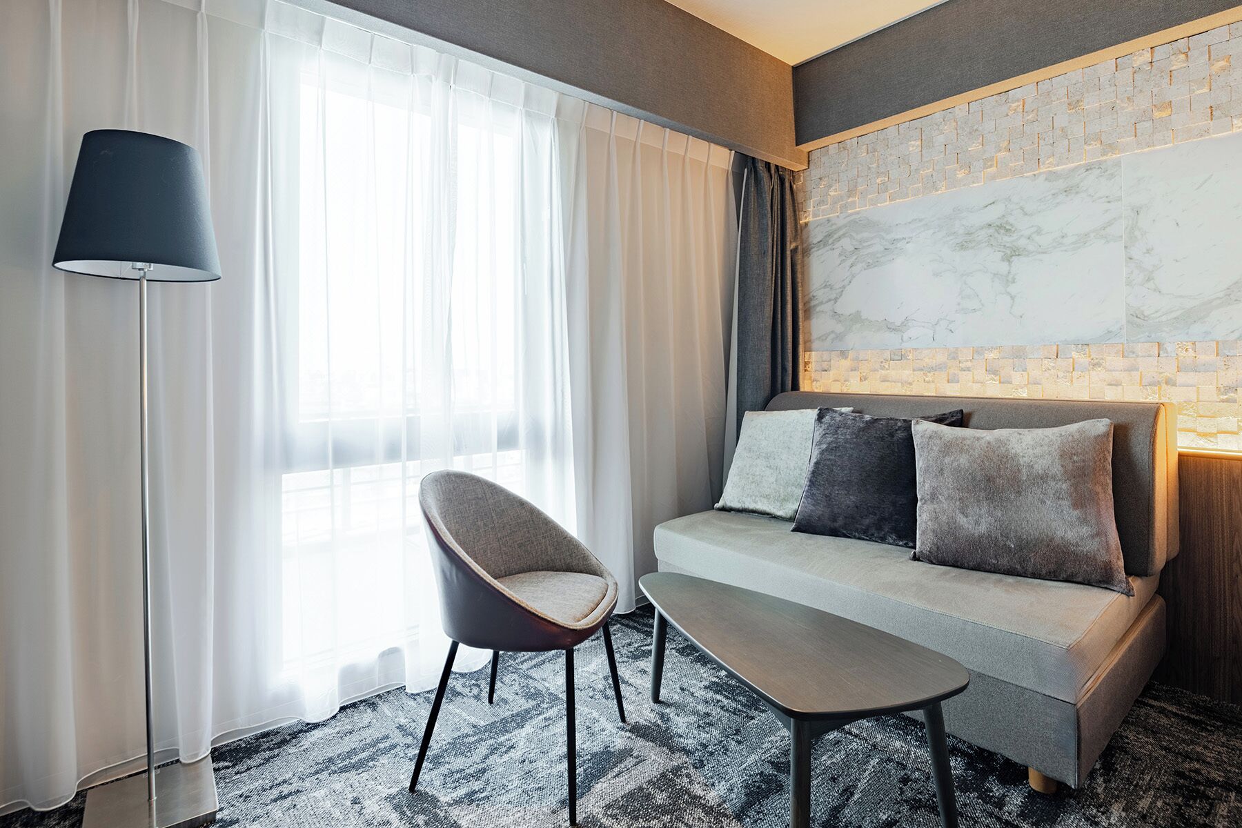 A modern hotel room with a sofa, a chair, a small table, and a lamp.