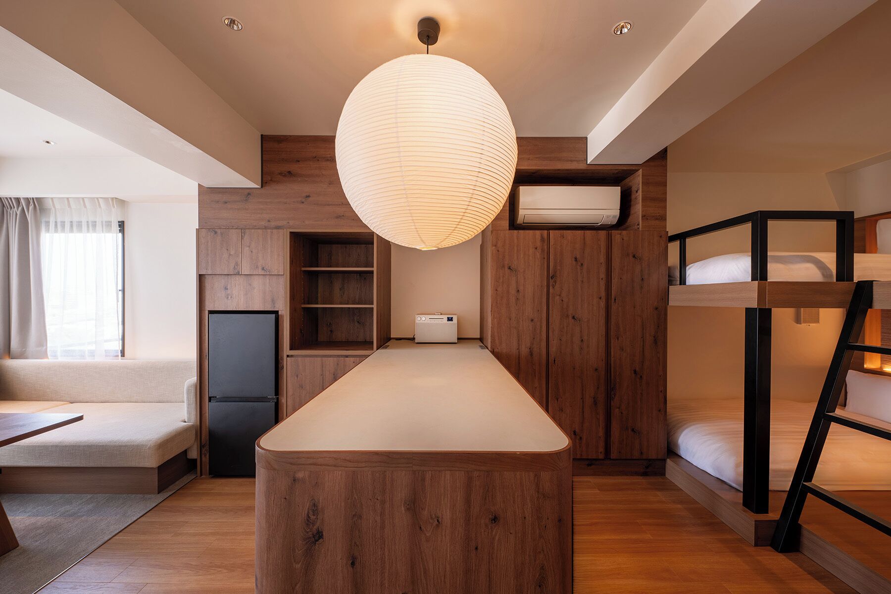 A compact hotel room with a wooden island table, bunk beds, a built-in radio, and a large hanging light fixture.