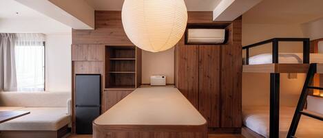 Quarto de hotel compacto com mesa ilha de madeira, beliches, rádio embutido e luminária pendente grande.
