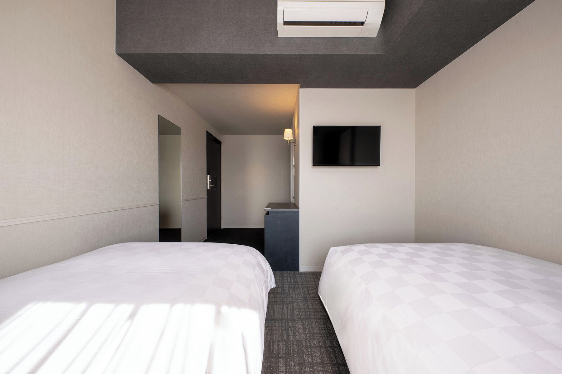 A hotel room with two beds, a flat-screen TV, a ceiling air conditioner, and a small desk with a lamp.