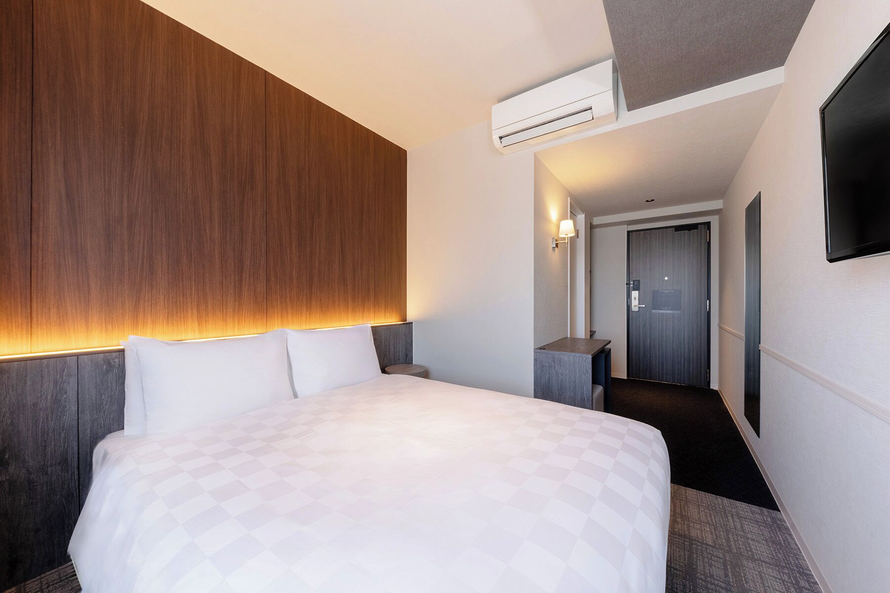 A hotel room with a large bed, a wooden headboard, an air conditioning unit, a flat-screen TV, and a hallway leading to a door.