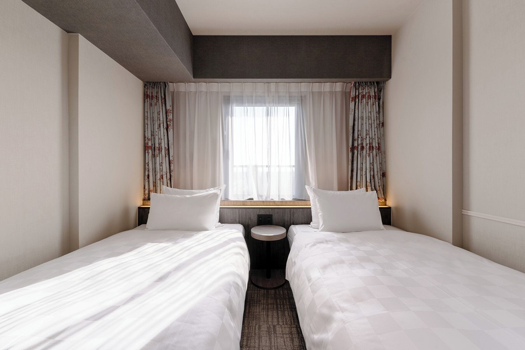 A hotel room with two single beds, a window with sheer curtains, and a small round table between the beds.