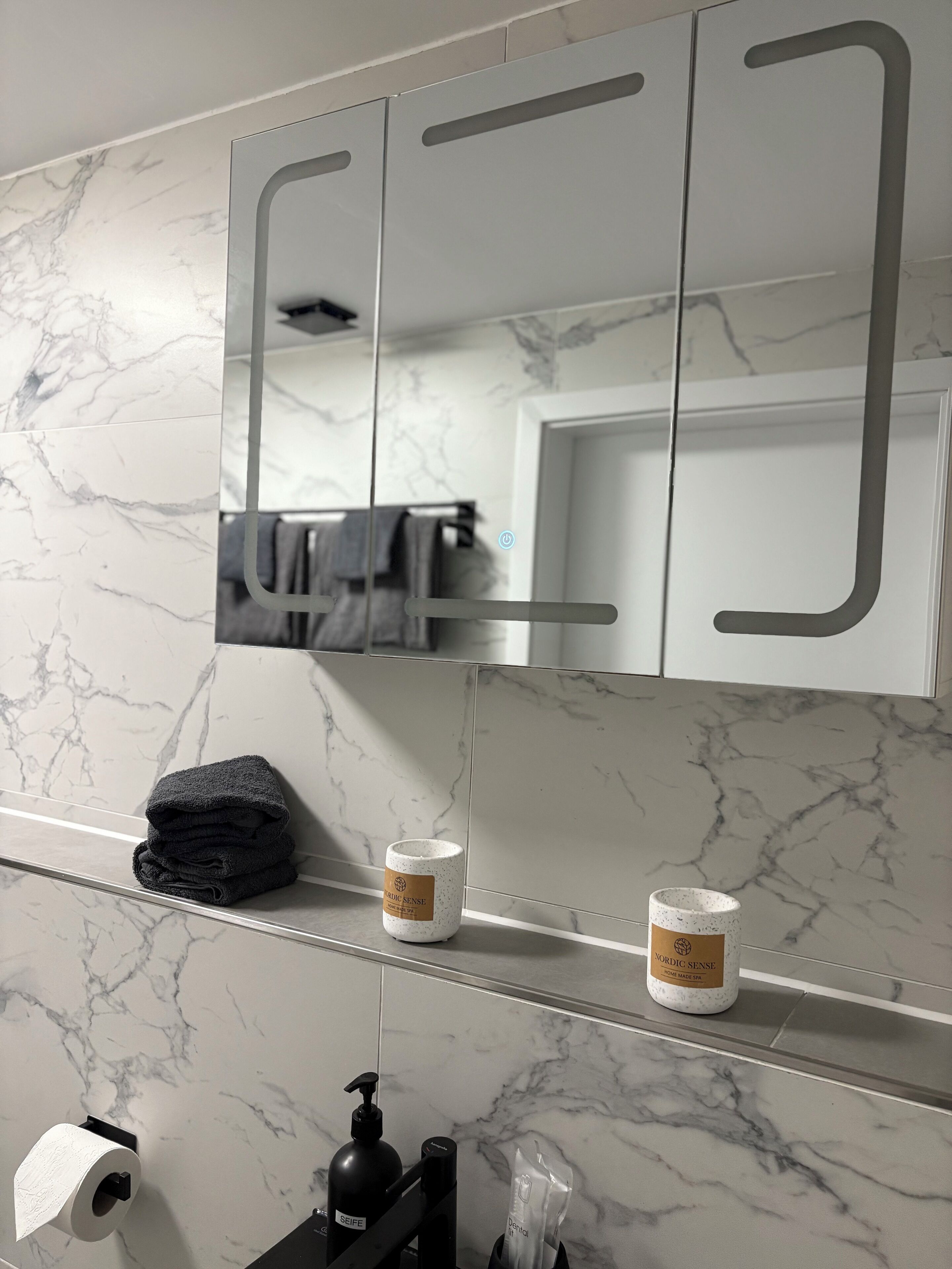 Une salle de bain avec un pare-feu en marbre, un miroir doté d’une poignée, une serviette et deux bouteilles étiquetées « Nordic Sense ».