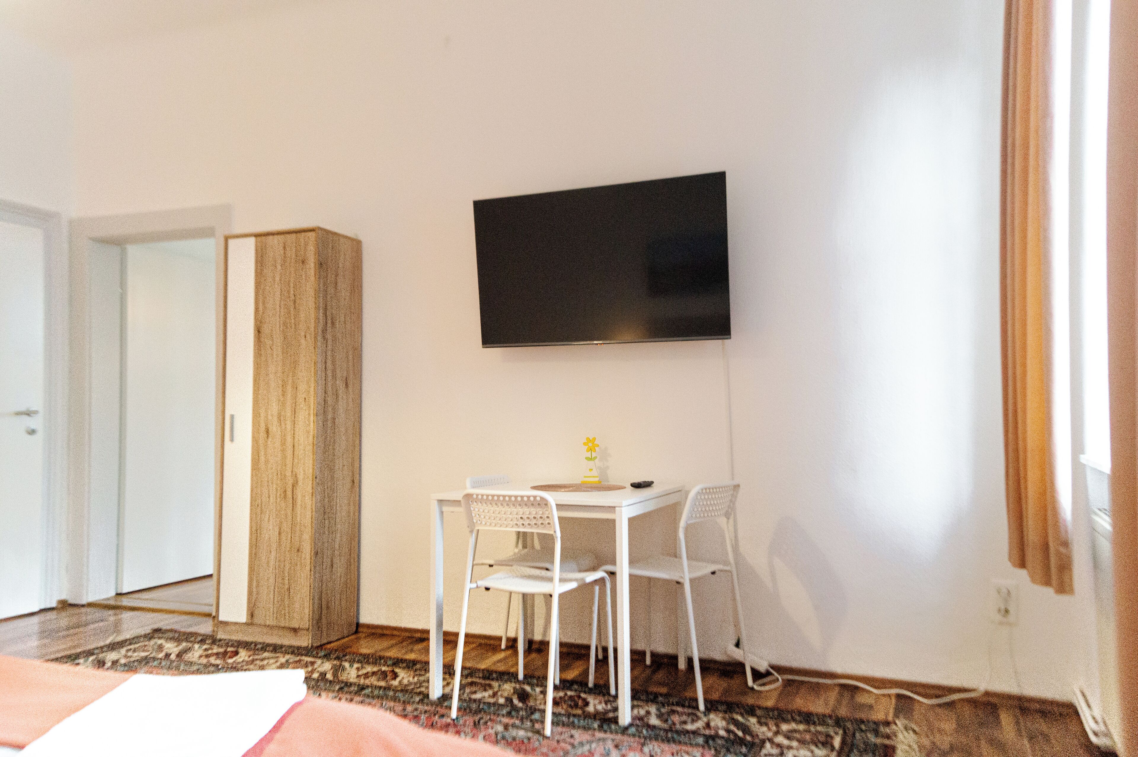 A room with a bed, a TV mounted on the wall, a wooden wardrobe, and a small table with two chairs.