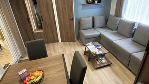 A compact living space with a sofa, a dining table with a basket of snacks, a coffee table with magazines, and a wall-mounted air conditioner. - At "Greg and Ilhem (Lège-Cap-Ferret)