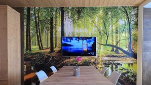 A modern dining area with a wooden table and chairs, a large TV screen displaying a forest scene, and a patterned rug on the floor. - Maison Cavalese - Wild Forest - Tranquility at 2 steps from the center (Cavalese)