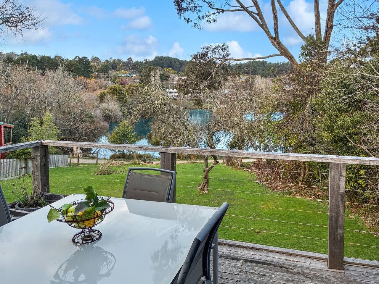 A dining table with a metal fruit basket on it, set against a backdrop of a grassy yard and a lake.