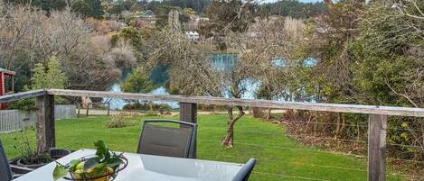 A dining table with a metal fruit basket on it, set against a backdrop of a grassy yard and a lake.