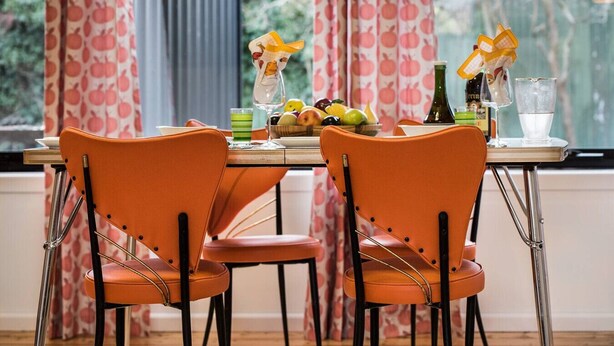 Een eethoek met twee oranje stoelen, een tafel met een fruitschaal, glazen en flessen.