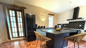 Una cocina moderna con una isla grande, dos taburetes de mimbre, un refrigerador negro y una pared con textura de piedra.