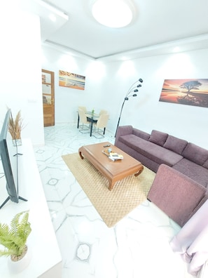 A modern living room with a sofa, coffee table, and TV. There is a dining area with chairs and a table in the background. - F3 air-conditioned with terrace - 1 minute from the beach (Dakar,Région de Dakar)