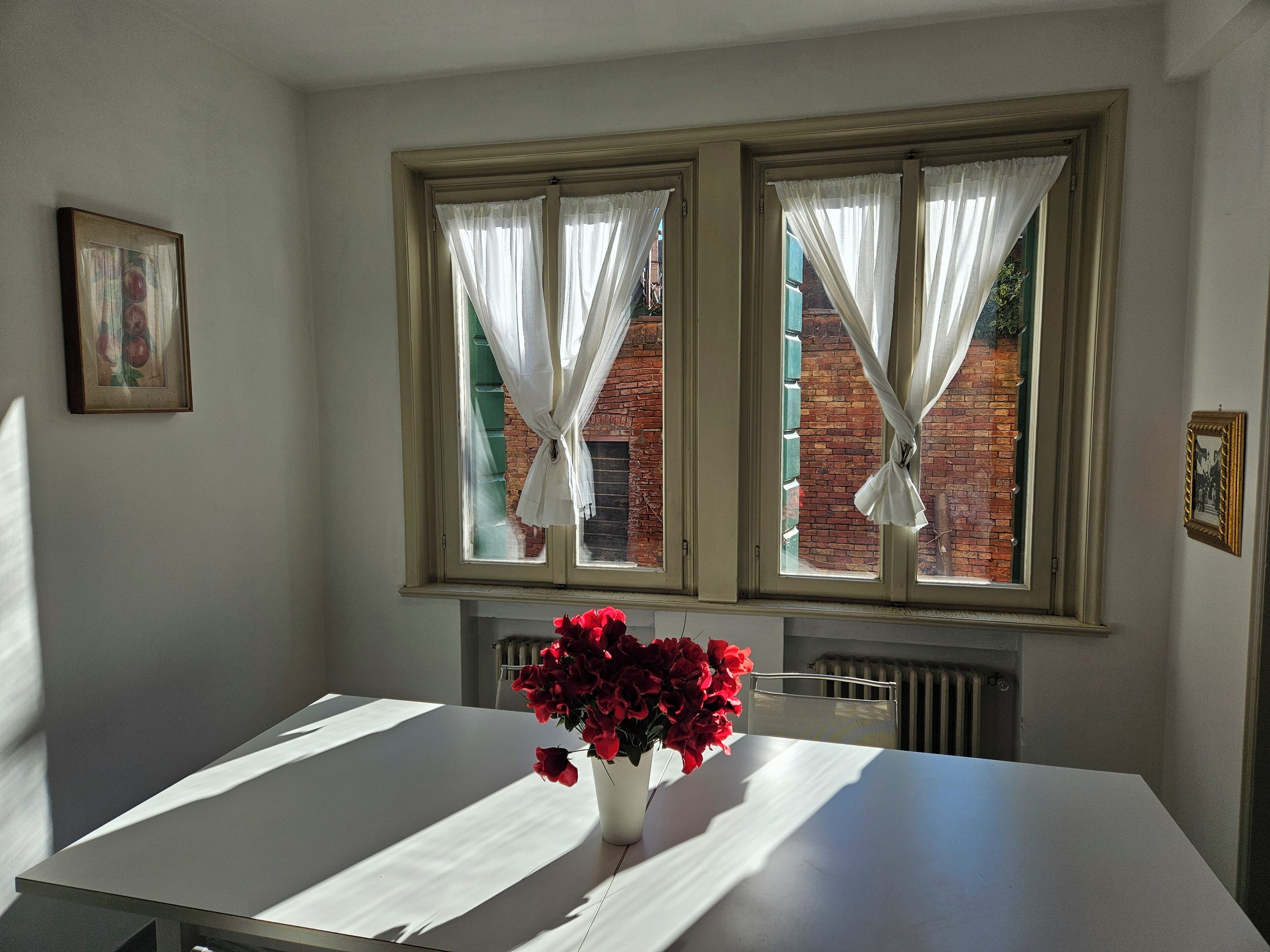 A room with a table, a vase with red flowers, and two windows with white curtains.