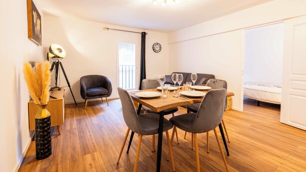 Un coin repas avec une table en bois dressée pour quatre personnes, entourée de chaises grises. Un lampadaire sur trépied et un vase contenant des plumes d’herbe des pampas se trouvent à gauche. Un canapé gris et un lit avec une couette blanche sont visibles à l’arrière-plan.