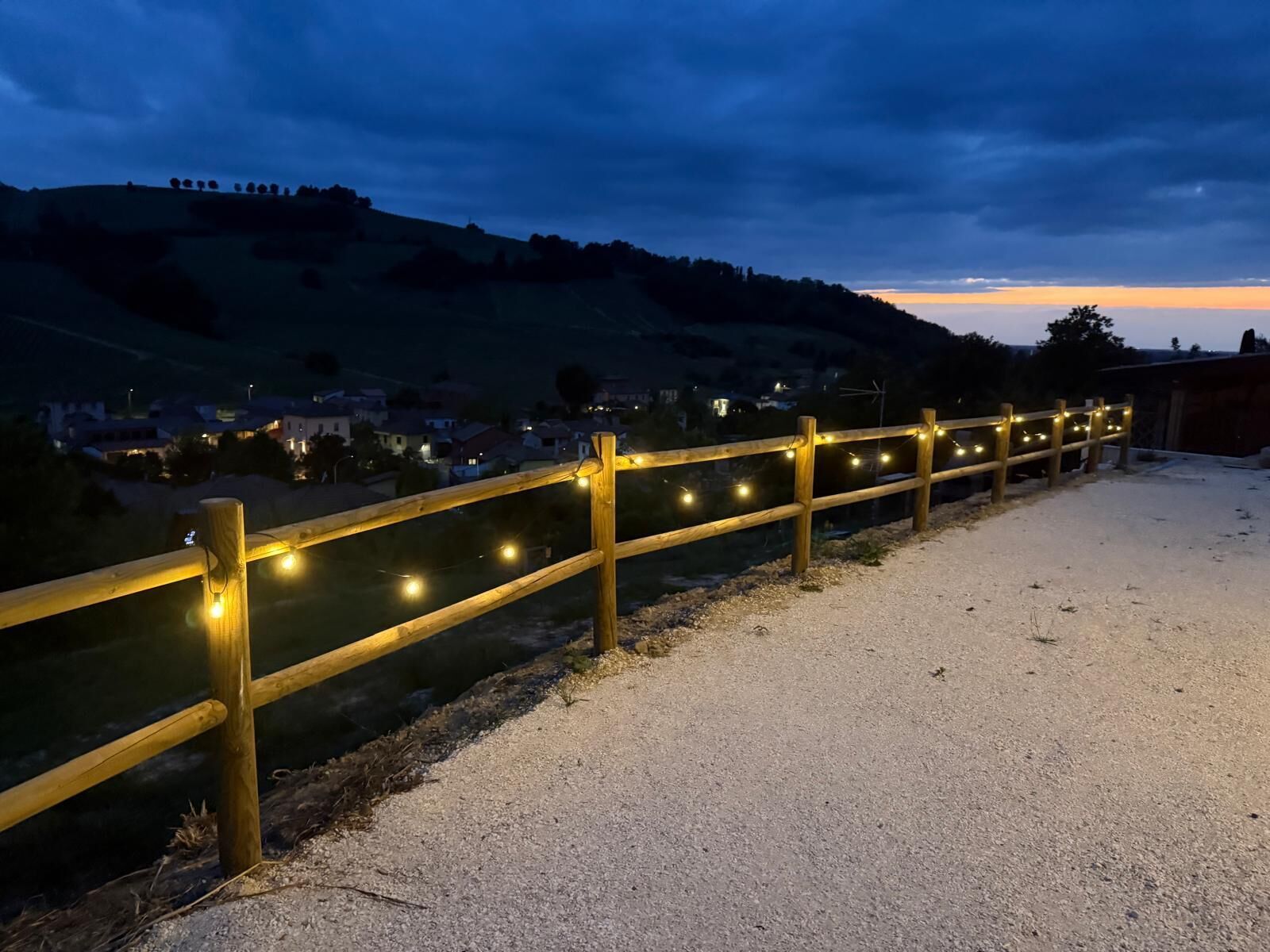 Un chemin de gravier bordé d’une clôture en bois et illuminé de guirlandes lumineuses, qui mène à une colline avec une ville en arrière-plan.