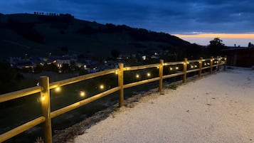Un chemin de gravier bordé d’une clôture en bois et illuminé de guirlandes lumineuses, qui mène à une colline avec une ville en arrière-plan.