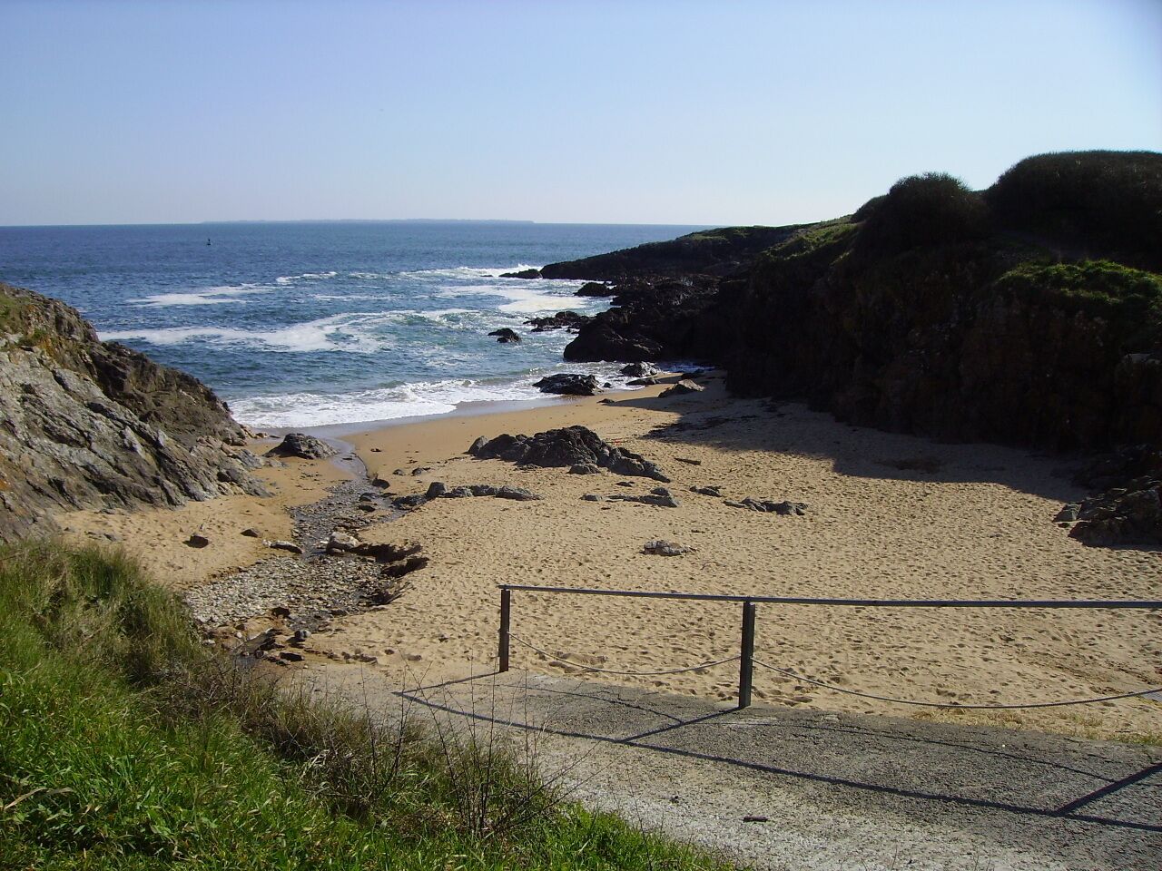 Ein Küstenstrand mit felsigem Ufer, ein sandiger Pfad, der zum Strand führt, und freie Sicht auf den Ozean.